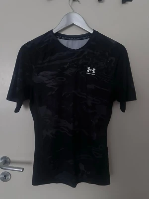 Svart compression-tshirt Under Armour - Säljer en svart tränings-tshirt från Under Armour med diskret mönster och logga på bröstet. T-shirten har rund hals och är tillverkad i ett lätt och snabbtorkande material som passar perfekt för intensiva träningspass.
