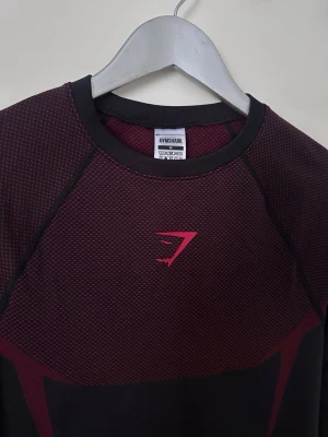 Gymshark onyx v5 - Snygg långärmad träningströja från Gymshark i burgundy och svart. Tröjan har ett tight fit, rund halsringning och mesh-liknande struktur upptill. Gymshark-logga i rosa på bröstet. Perfekt för gymmet eller löpning.