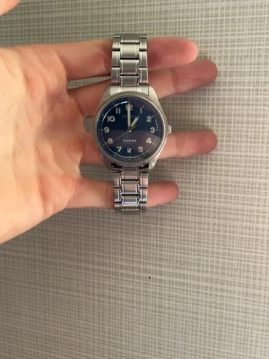 Blå analog klocka med metallarmband - Snygg analog klocka från Timex med blå urtavla och tydliga siffror. Klockan har ett klassiskt silverfärgat metallarmband och datumvisning. Perfekt för dig som gillar stilrena accessoarer med en modern touch.