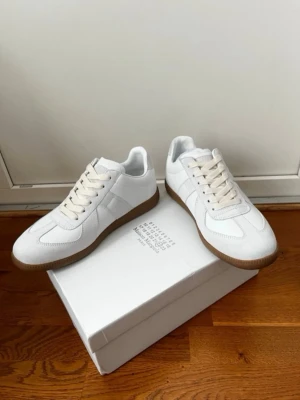  Maison Margiela - Maison Margiela sneakers i vitt läder och mocka med klassisk design och beige gummisula. Skorna har vita snören och diskreta paneler i olika material som ger en snygg kontrast. Perfekt för dig som gillar stilrena och trendiga sneakers.