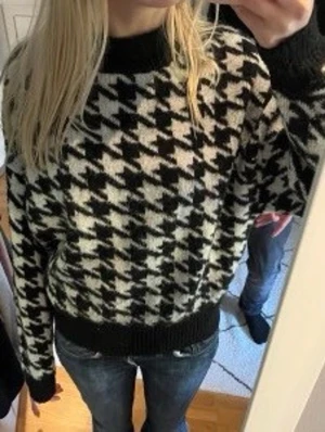 Svartvit stickad tröja med hundtandsmönster - Cool stickad tröja med klassiskt svartvitt hundtandsmönster. Tröjan har rund halsringning och ribbade muddar vid ärmslut och nederkant. (Köpt second hand) 