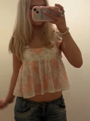 Blommig volang topp med breda axelband - Söt blommig topp med volang och breda axelband. Toppen är i ljusa färger med rosa och orange blommor på vit botten. Luftigt material, perfekt för varma dagar. Axelbanden har volangdetaljer som ger en romantisk vibe.