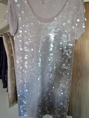GUESS/ Silver paljett-topp med kort ärm - Snygg silverfärgad lång topp med korta ärmar och rund halsringning. Kan öven användas som kort klänning.Framsidan är täckt av glittrande paljetter som ger ett riktigt coolt och festligt uttryck. Baksidan är enkel och slät. Perfekt för dig som vill sticka ut. Mycket bra skick.