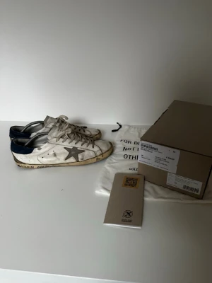 Golden Goose  - Golden Goose sneakers i vitt läder med den ikoniska grå stjärnan på sidan och mörkblå häl. Skorna har vita snören, blå yttersula och detaljerade sömmar. Medföljer originalkartong och dustbag. Perfekta för dig som gillar en avslappnad och trendig stil. Allt og ingår! 