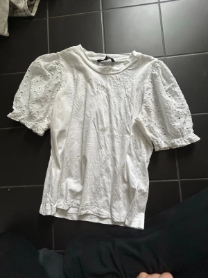 Vit blus med puffärm och spets - Säljer en vit blus med puffiga kortärmar i spets och broderade detaljer. Blusen har rund halsringning och är gjord i bomull. Passar underbart på en sommarkväll eller bara en dag i skolan! 
