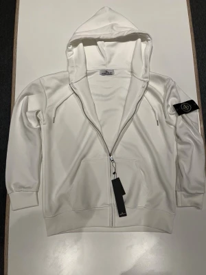 Stone Island zip hoodie – vit - Säljer en vit Stone Island zip hoodie i nyskick, helt oanvänd med originaltags kvar. Klassisk modell med dragkedja, huva och den ikoniska Stone Island-kompassen på ärmen. Perfekt för både vardag och stilren look.