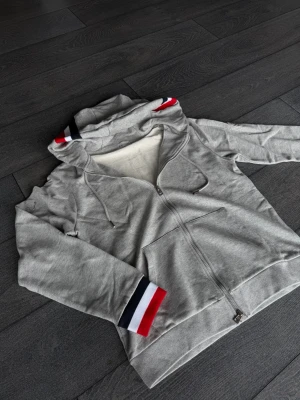 Grå moncler zip up - Snygg grå hoodie med dragkedja och huva. Ärmsluten och huvan har detaljer i rött, vitt och blått för en sportig vibe. Hoodien har fickor framtill och är gjord i mjukt sweatshirtmaterial, perfekt för chill dagar eller streetwear-stil.