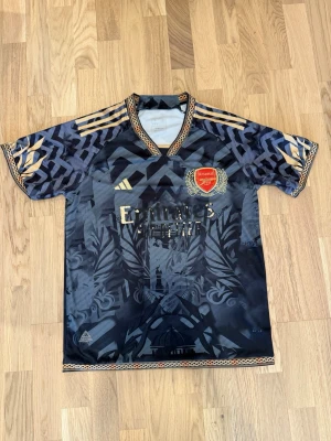 Arsenal tröja - Snygg Arsenal tröja från Adidas med guldiga detaljer och klubbmärke på bröstet. Tröjan har ett unikt mönster, V-ringad krage och tre guldiga ränder på axlarna. Tillverkad i lätt och ventilerande material, perfekt för fotboll eller träning. Storlek L men passar som M och S, jag är 170cm och 72kg och ni kan se hur den sitter på mig.