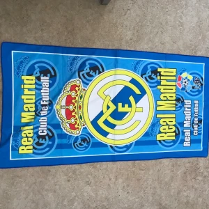 Real Madrid strandhandduk blå - Cool strandhandduk med Real Madrid-logga och klubbnamn i gult på blå bakgrund. Handduken har stora klubbemblemet i mitten och detaljer med fotbollsmotiv. Perfekt för stranden eller poolen för dig som älskar fotboll och Real Madrid.