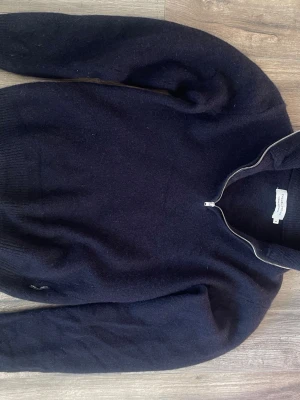 Mörkblå halvzip stickad tröja - Mörkblå stickad tröja från Oscar Jacobson med halvzip och hög krage. Tröjan har långa ärmar och ribbstickade muddar vid ärmslut och nederkant. Materialet är mjukt och värmande, perfekt för kyliga dagar. Pris inte hugget i sten så kom med pris förslag