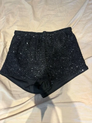 Svarta paljettshorts - Glittriga svarta shorts täckta med paljetter. De har en hög midja och är tillverkade i ett stretchigt syntetmaterial. Perfekta för att sticka ut och skapa en cool look.