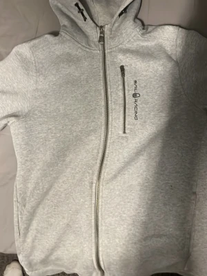 Ljusgrå hoodie från Sail Racing - Ljusgrå hoodie från Sail Racing med hel dragkedja framtill och en extra bröstficka med dragkedja. Hoodien har en broderad logga på bröstet och en mindre logga på huvan. Tillverkad i mjukt material och har en sportig, clean look. Passar inte mig längre vill bli av med den snabbt. Passar både killar och tjejer med storleken S  
