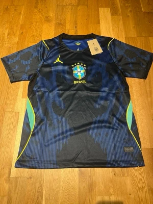 Blå Brasilien matchtröja Nike Jordan - Snygg blå och svart Brasilien matchtröja från Nike Jordan med gula detaljer och CBF-logga på bröstet. Tröjan har korta ärmar, rund hals och är tillverkad i ett lätt, ventilerande funktionsmaterial. Perfekt för fotboll eller träning.
