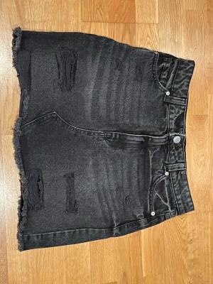 Svart jeanskjol med slitningar - Säljer en svart kort jeanskjol med slitna detaljer och rå kant nertill. Kjolen har klassisk femficksdesign och knappstängning fram. Perfekt för dig som gillar en edgy och avslappnad stil.