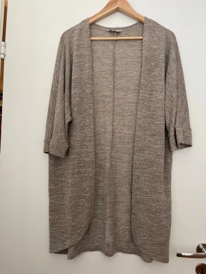 Beige lång kofta med uppvikta ärmar - Mysig beige lång kofta i mjukt stickat material. Koftan har öppet fall, uppvikta trekvartsärmar och en avslappnad passform. Perfekt att slänga över en t-shirt eller topp för en chill look.