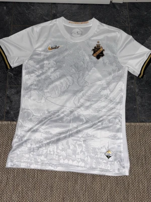 Vit AIK matchtröja från Nike - Säljer en vit AIK matchtröja från Nike med snyggt mönster av Stockholm på framsidan. Tröjan har AIK:s klubbmärke i svart och guld på bröstet, guldfärgade detaljer och svarta/guldiga ränder på ärmsluten. Perfekt för dig som vill representera AIK på läktaren eller träningen.
