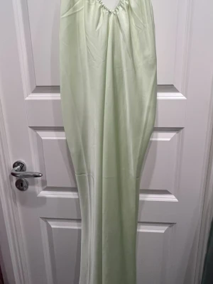 Ljusgrön långklänning från Lindex - Långklänning i ljusgrön satin från Lindex. Klänningen har halterneck med knytband i nacken och ett stilrent, elegant fall. Ärmlös design och loose passning som ger en skön och glansig look.