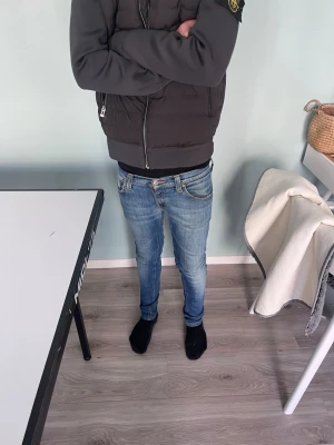Blå slim fit jeans från Nudie Jeans - Säljer ett par blå slim fit jeans från Nudie Jeans med klassisk femficksmodell och snygga slitningar. Jeansen har kontrastsömmar och bruna detaljer på bakfickorna. Perfekta för dig som gillar en smal passform och tidlös stil.