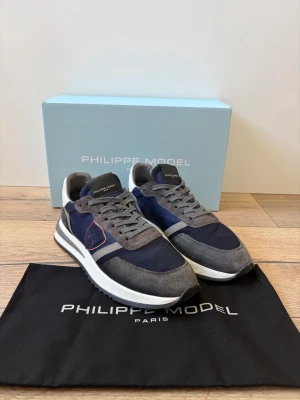 Philippe Model blå/grå sneakers - Snygga sneakers från Philippe Model i blått och grått med mesh och mockadetaljer. Skorna har vita och svarta inslag på sulan, klassisk låg profil och logotyp på sidan. Perfekta för dig som vill ha en stilren och trendig look.