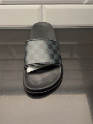 Svart-rutig badtoffla - Säljer en svart badtoffla med klassiskt rutigt mönster i grått och svart på remmen. Tofflan har en bred rem över foten och en bekväm, formad sula i syntetmaterial. Perfekt för chill dagar hemma eller vid poolen.