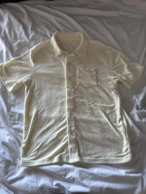Beige frottéskjorta med kort ärm - Säljer en beige skjorta i frottématerial med klassisk krage, korta ärmar och bröstficka. Skjortan har knappar framtill och en avslappnad passform som är perfekt för en chill look.