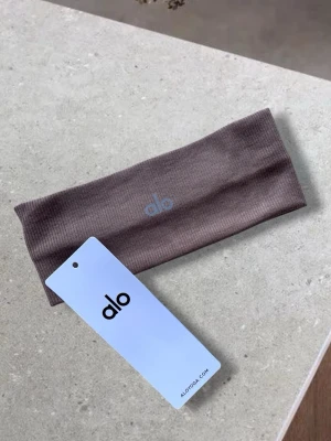 ALO Ribbat ljusbrunt hårband  - Säljer ett ribbat hårband i ljusbrun färg från Alo. Hårbandet är brett och elastiskt, vilket gör det bekvämt och lätt att använda till olika frisyrer. Materialet är mjukt och stretchigt för en skön känsla hela dagen.