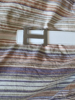 Vitt skärp med H-spänne - Säljer ett stilrent vitt skärp med ett stort H-format spänne i silverfärgad metall. Skärpet är gjort av skinn och har tydliga sömmar längs kanterna. Perfekt för att lyfta din outfit med en modern touch.