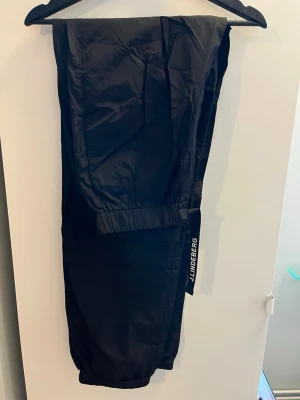 Svarta byxor från J.Lindeberg - Säljer ett par svarta byxor från J.Lindeberg med elastisk midja och justerbart bälte. Byxorna har en sportig look och är tillverkade i ett glansigt, vindtätt material. Perfekta för kalla dagar och outdoor-aktiviteter.