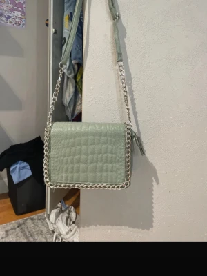 Ljusgrön kedjedetaljerad handväska - Säljer en ljusgrön handväska med quiltad design och silverfärgad kedja runt kanterna. Väskan har justerbar axelrem och flera fack inuti, inklusive ett med dragkedja. Materialet är konstläder och insidan är brun textil. Perfekt för dig som gillar stilrena accessoarer.