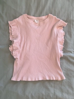 Ljusrosa ribbad topp med volang - Supersöt ljusrosa topp från Zara i ribbad bomull med volangdetaljer längs ärmarna. Modellen är ärmlös med rund halsringning och har en feminin, mjuk look som passar perfekt till jeans eller kjol.