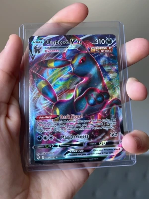 Pokémonkort Umbreon VMAX Full Art - Säljer ett Pokémonkort Umbreon VMAX Full Art med holografisk yta. Kortet har färgstarka detaljer och motiv av Umbreon, med text och symboler i silver och guld. Kortet är inplastat i ett skyddsfodral och har en unik design från VMAX-serien. Den är väldigt sällsynt och mycket bra för samlare inom pokemon