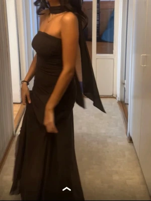 Mörkbrun långklänning med scarf - Elegant mörkbrun långklänning med axelbandslös design och matchande scarf runt halsen. Klänningen är slutsåld på Nelly och är endast använd en gång.