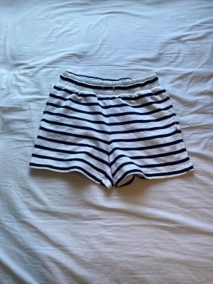Randiga shorts från Lindex - Snygga vita shorts med mörkblå ränder från Lindex. De har resår i midjan för en bekväm passform och är perfekta för varma dagar. Tillverkade i mjuk bomull som känns skön mot huden.
