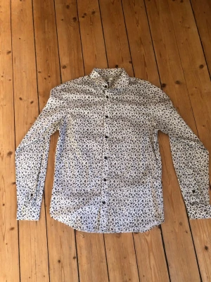 Vit blommig skjorta med knappar - Snygg vit skjorta med små blommiga mönster i blått och svart. Skjortan har lång ärm, klassisk krage och knäppning framtill. Perfekt för dig som vill sticka ut lite extra med ett coolt print.