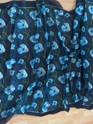 Blommig scarf i blått och svart - Snygg besch scarf med transparent svart bas och stora blå blommor med gröna stjälkar. Materialet är tunt och lätt, perfekt att slänga över axlarna för en cool look. Mönstret är livligt och färgstarkt, vilket gör sjalen till ett statement-piece.