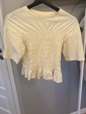 Ljus gul T-shirt med ribbning från Gina  - Ljus gul T-shirt smockad midja och volangkant från Gina. Den är i storlek S men passar även XS. Använd fåtal gånger 