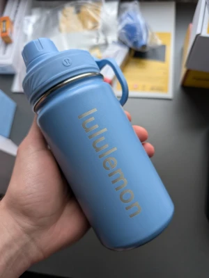 Blå vattenflaska från Lululemon - Snygg blå vattenflaska från Lululemon med praktiskt skruvlock och handtag. Flaskan är tillverkad i rostfritt stål och har Lululemon-loggan tryckt i silver på sidan. Perfekt för att ta med till gymmet eller skolan.