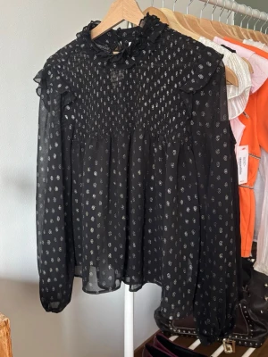 Svart blus med silverprickar från Nelly  - Svart blus från Nelly med silverfärgade prickar och smockad överdel. Blusen har volangdetaljer vid halsen och axlarna samt långa ärmar i transparent material. 