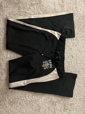 Svarta träningsshorts från Max Azria - Snygga svarta byxor från Max Azria med silverfärgade detaljer längs sidorna och tryckt emblem på ena benet. Byxorna har snörning i midjan och är tillverkade i ett lätt och sportigt material som passar perfekt för aktiva dagar.