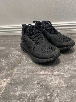 Svarta Nike Air Max 270 sneakers - Säljer ett par helsvarta Nike Air Max 270 sneakers med mesh-överdel och synlig Air-enhet i hälen. Skorna har en sportig look med klassisk snörning och diskreta detaljer. Perfekta för dig som gillar streetwear och vill ha bekväma sneakers med modern design.