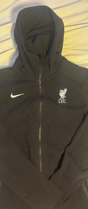 Svart Nike Liverpool hoodie - Säljer en svart hoodie från Nike med Liverpool FC-logga på bröstet. Hoodien har dragkedja, huva och två fickor med dragkedja. Storleken är s men själv brukar jag köra M och är 187 lång.