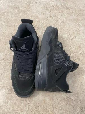 Svarta Air Jordan 4 sneakers - Säljer ett par svarta Air Jordan 4 sneakers med klassisk Jumpman-logga på plösen. Skorna har meshdetaljer, robust gummisula med synlig Air-enhet och coola plastdetaljer vid snörningen. Perfekt för dig som vill ha en ikonisk och stilren sneaker.