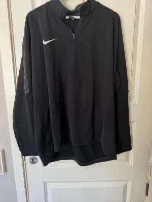 Svart Nike Dri-Fit träningströja - Säljer en svart långärmad träningströja från Nike med Dri-Fit material som håller dig torr under träning. Tröjan har halv dragkedja framtill och diskret Nike-logga på bröstet. Perfekt för sport och aktivitet.