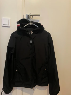 Svart vindjacka med huva - Snygg svart vindjacka med huva och dragkedja framtill. Jackan har två fickor med tryckknappar och en detalj med röd och vit rand på huvan. Perfekt för blåsiga dagar och enkel att matcha med olika outfits.