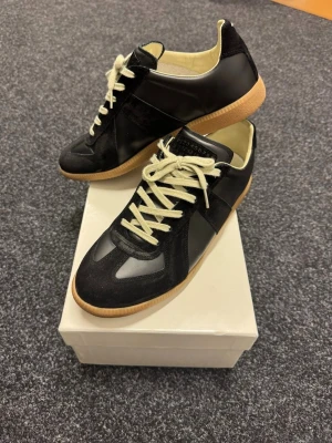 Maison Margiela GATS - Maison Margiela sneakers i svart läder och mocka med beige gummisula och vita snören. Klassisk German Army Trainer-design med låg profil och stilren look. Perfekt för dig som vill ha en tidlös och trendig sneaker.