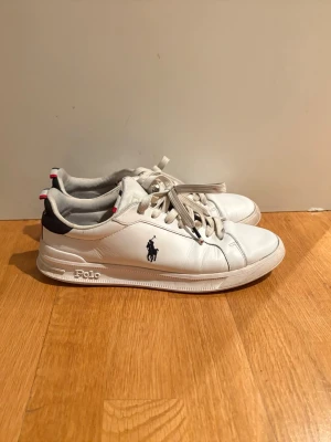Vita Polo Ralph Lauren sneakers - Snygga vita sneakers från Polo Ralph Lauren med klassisk logga på sidan och guldtext på hälen. Skorna har mörkblå detaljer och röda/vita ränder bak. Tillverkade i läder med textilfoder och platt sula. Perfekta för en stilren look.