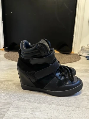 Svarta wedge sneakers med kardborre - Säljer ett par svarta sneakers med hög kilklack och kardborreband. Skorna har en mix av läder och textilmaterial, samt snörning framtill. De har en cool och modern design med högt skaft och detaljer i olika texturer. Perfekta för dig som vill ha både stil och attityd.