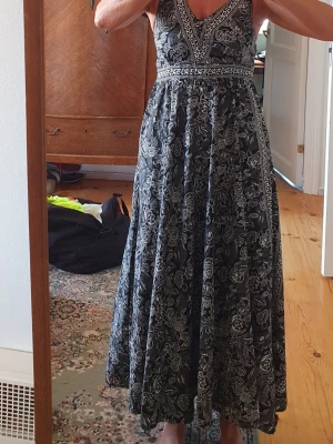 Svart långklänning med blommigt mönster - Svart långklänning med vitt blommigt mönster och markerad midja. Klänningen har v-ringning och är ärmlös, med ett luftigt och elegant fall. Perfekt för dig som gillar bohemisk stil och vill ha något bekvämt men snyggt.