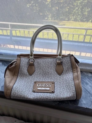 Beige och vit handväska från Guess - Snygg handväska från Guess i beige och vitt med logomönster och stora handtag. Väskan har ett stort fack med dragkedja och leopardmönstrat innerfoder. Den har varit väl omhändertagen och använt på ett bra sätt. Inge effekter förekommer, den har tre fack. Passar dig som vill ha en medelstor väska och mönster på insidan. 
