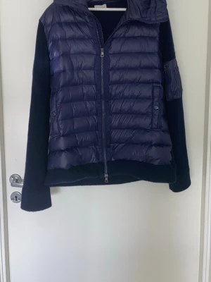 Blå dunväst med huva från Moncler - Snygg blå dunväst från Moncler med stickade ärmar och huva. Västen har en quiltad front i glansigt material, dragkedja och Moncler-logga på ärmen. Perfekt för lager-på-lager och streetstyle. Passar dig som vill ha både värme och stil. Pris kan diskuteras. 